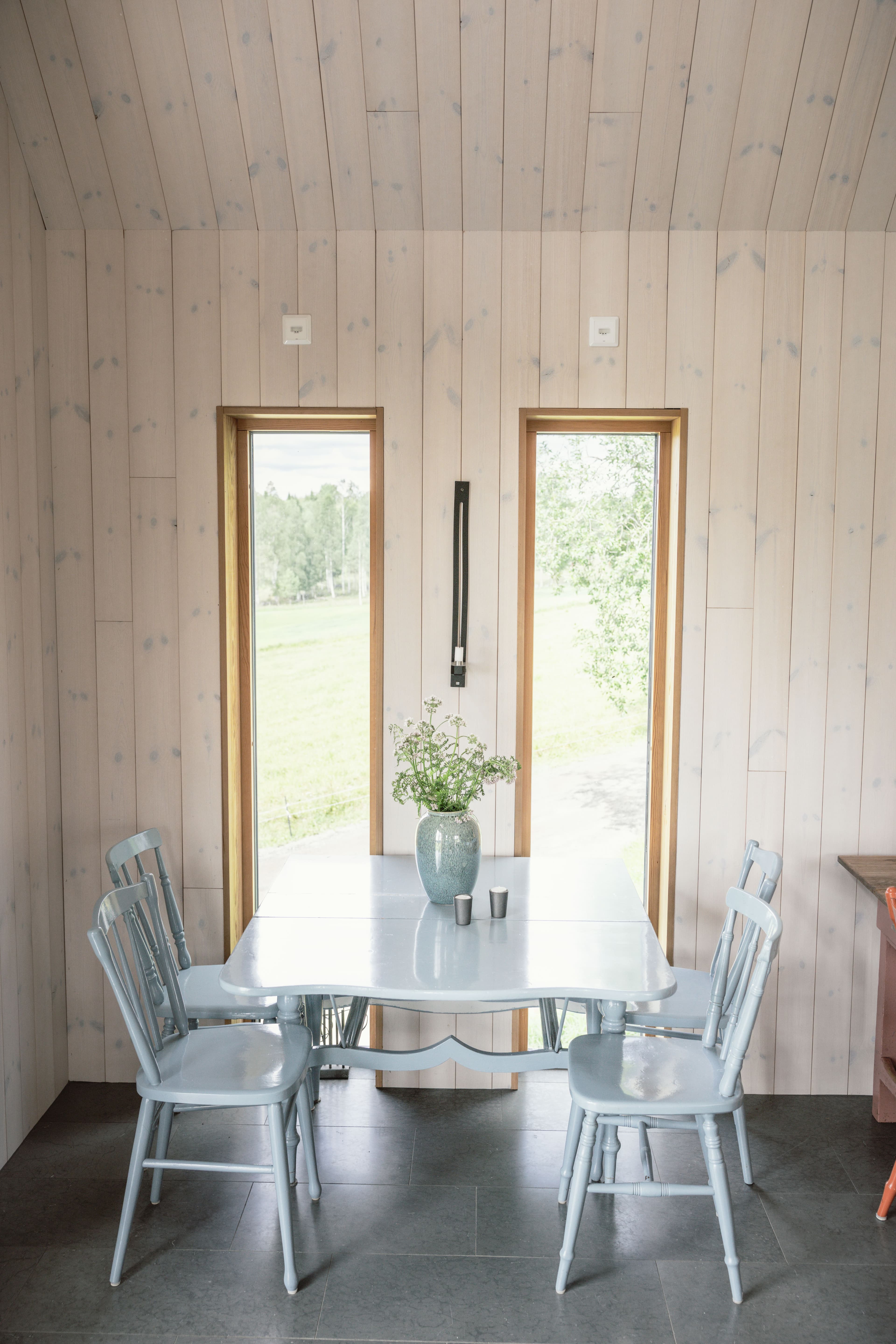 Matbord med, fyra ljusblå stolar och en vas med blommor. Två höga fönster mot grönskande landskap, väggpanel från Norrlands trä, Putsad slätspont Superslasyr vit.