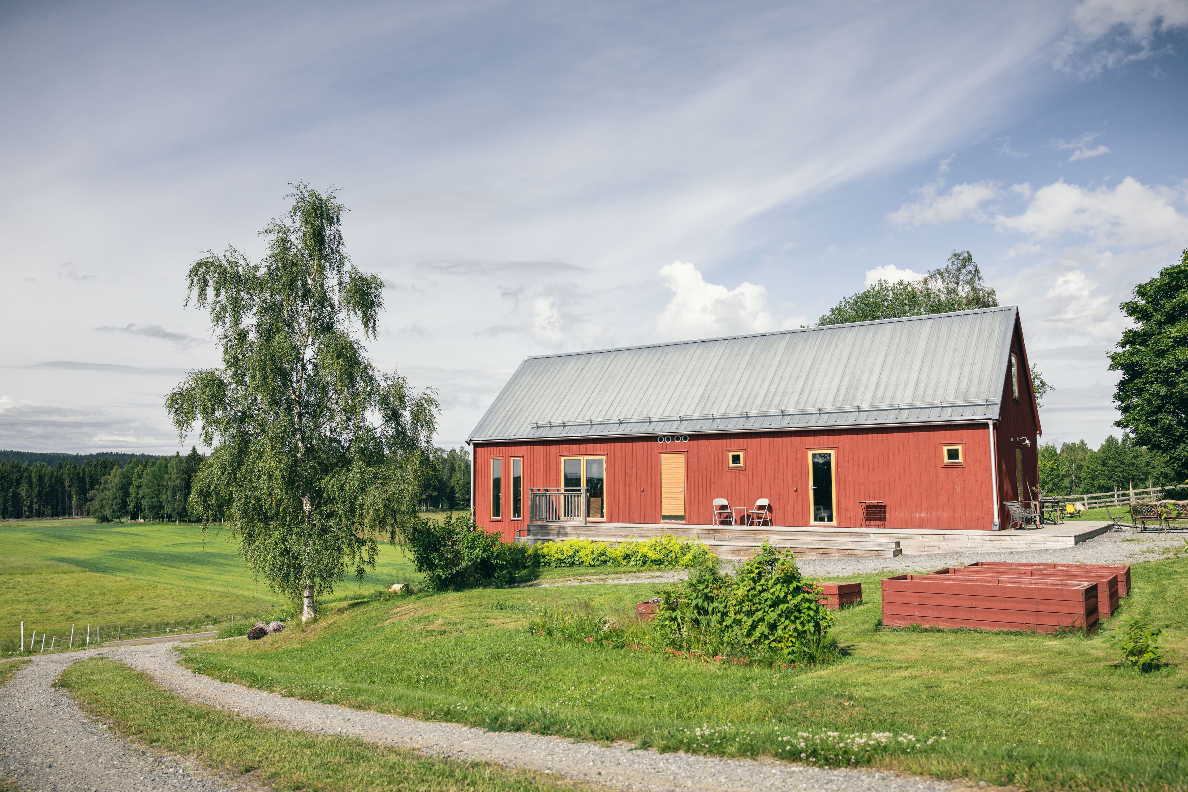 Boda Gård, ett rött ladugårdsliknande hus, omgivet av gröna ängar och träd, under en delvis molnig himmel.
