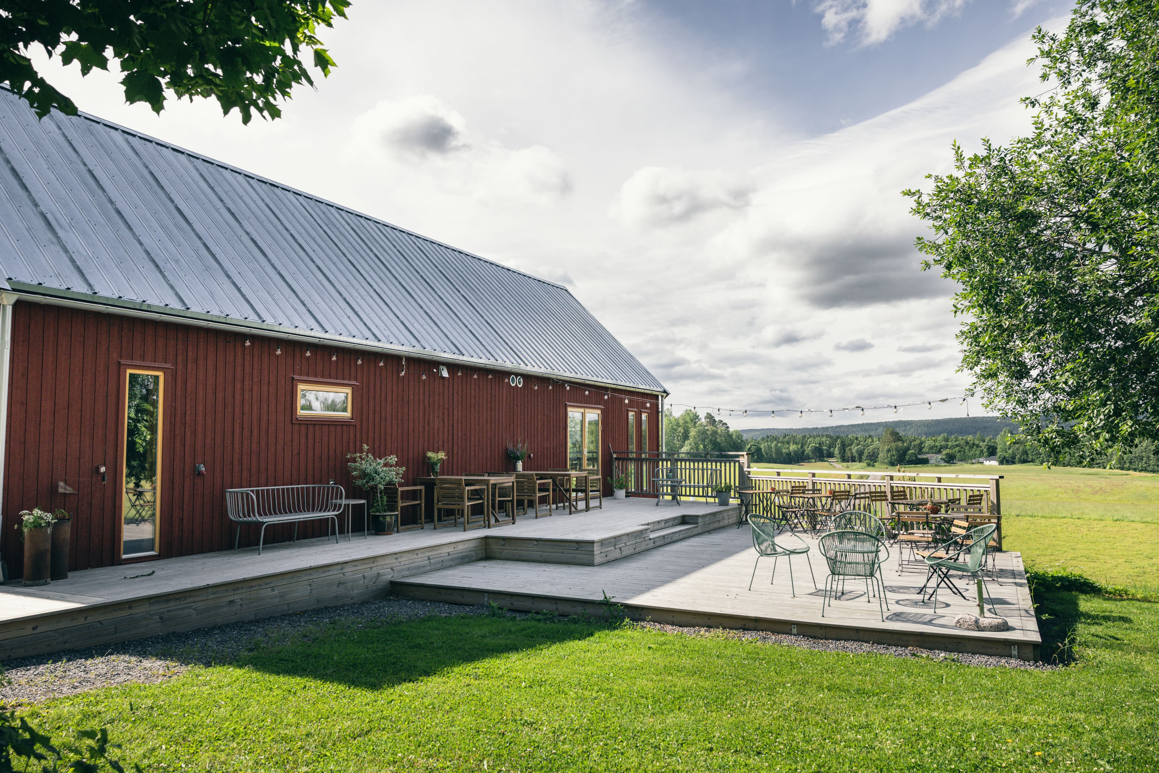 En röd lada med grått plåttak under en delvis molnig himmel. Framför ladan breder en rymlig träaltan ut sig, prydd med utemöbler.