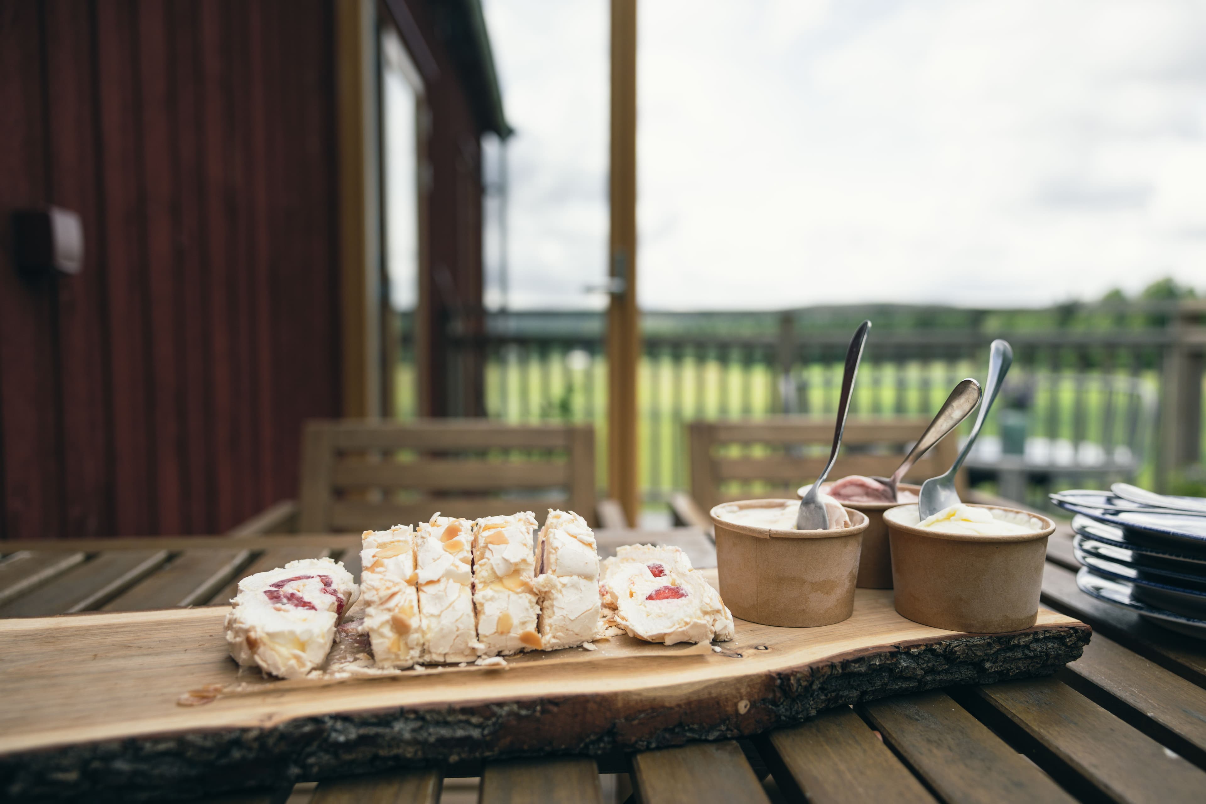 Träbricka med rulltårtor och tre papperskoppar med egengjord glass. Utomhus på ett altanbord.